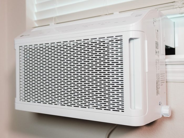 White window air conditioner installed in a room with closed blinds and power outlet visible on the wall