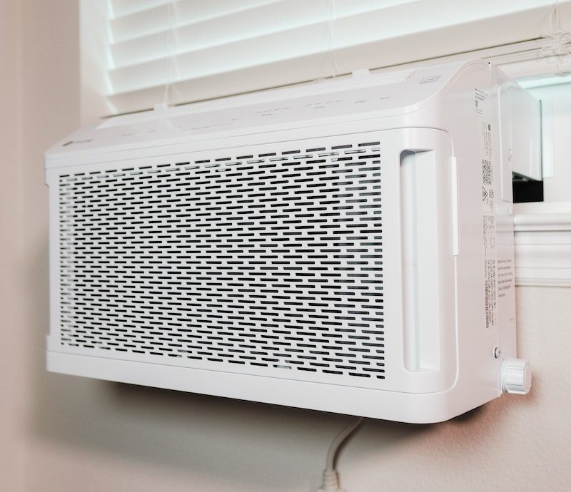 White window air conditioner installed in a room with closed blinds and power outlet visible on the wall