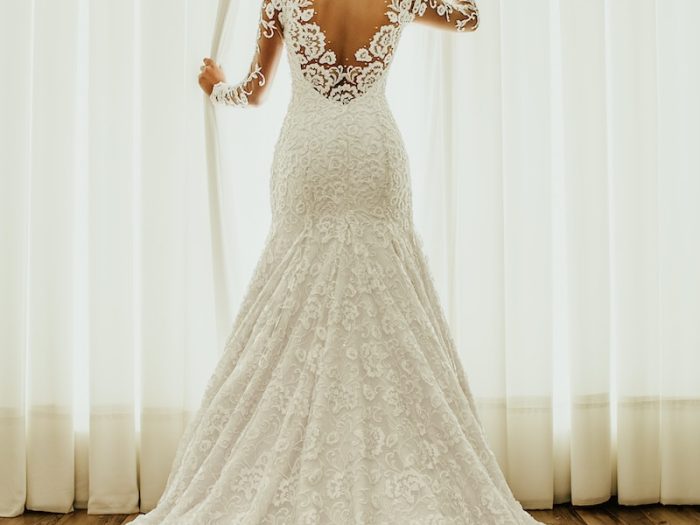 Bride in elegant lace wedding dress stands before bright window curtains on polished wooden floor