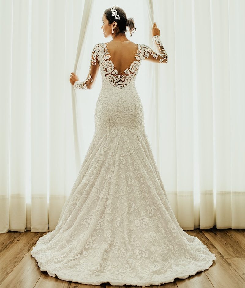 Bride in elegant lace wedding dress stands before bright window curtains on polished wooden floor