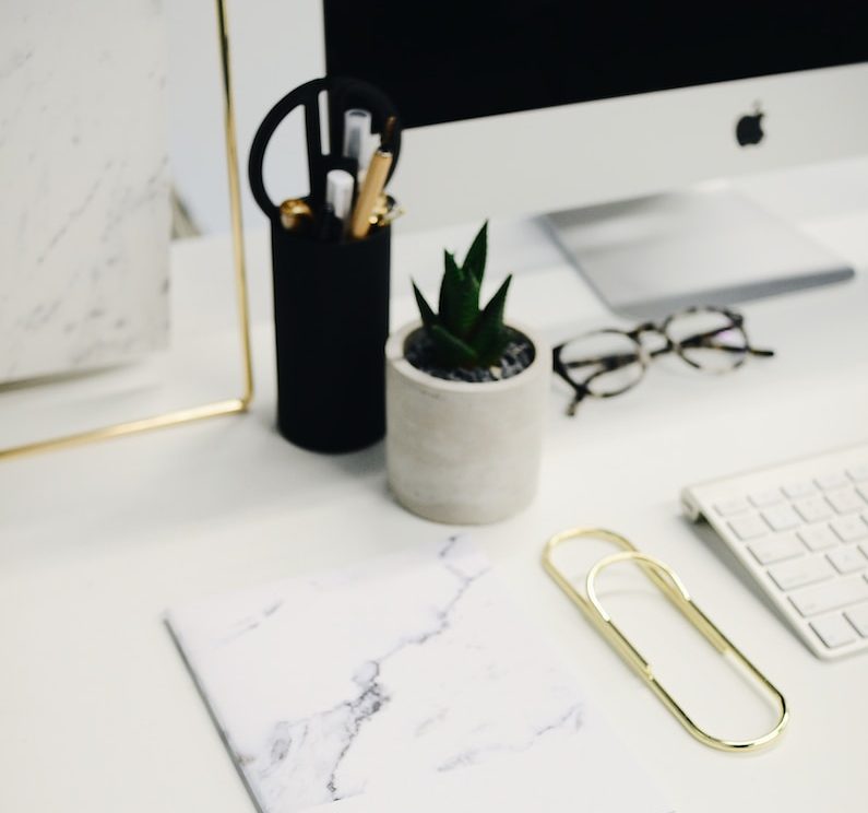 Sleek office desk setup with iMac, keyboard, glasses, notebook, pen holder, plant, and gold paperclip on a white surface
