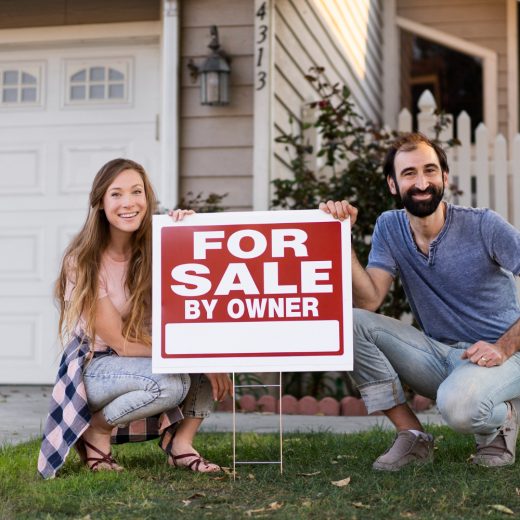A couple stands together, holding a "For Sale by Owner" sign in front of their house, smiling at the camera