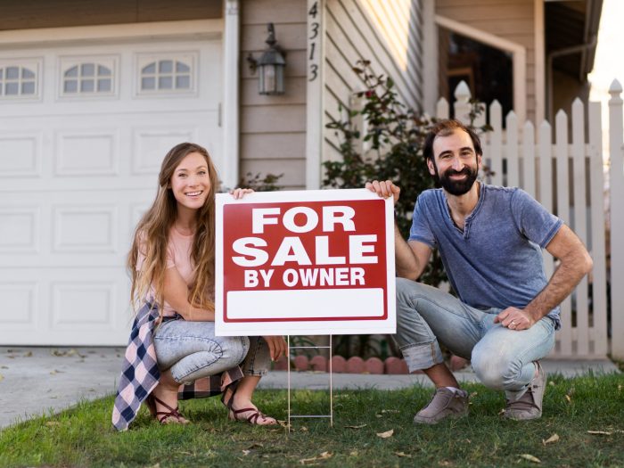 A couple stands together, holding a "For Sale by Owner" sign in front of their house, smiling at the camera